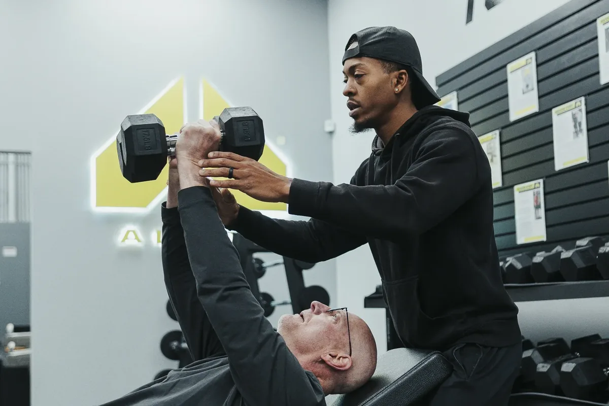 Alloy coach guiding a member through a dumbbell press during a personal training session