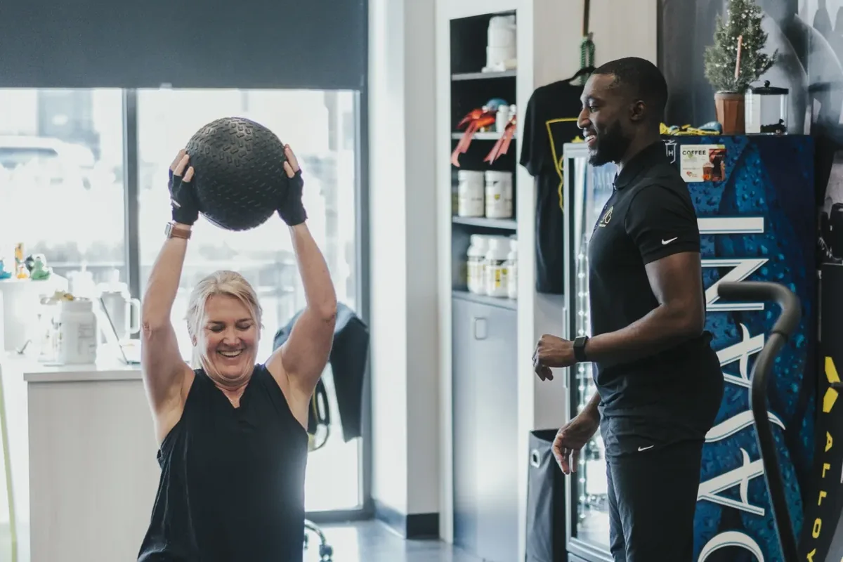 A woman presses a medicine ball overhead from a kneeling position while an Alloy coach looks on in the studio