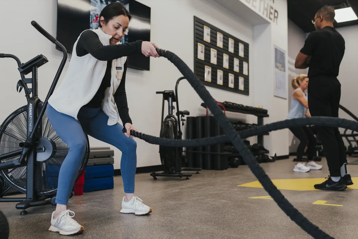 A woman works battle ropes while a coach looks on during a small group training session at an Alloy Personal Training studio