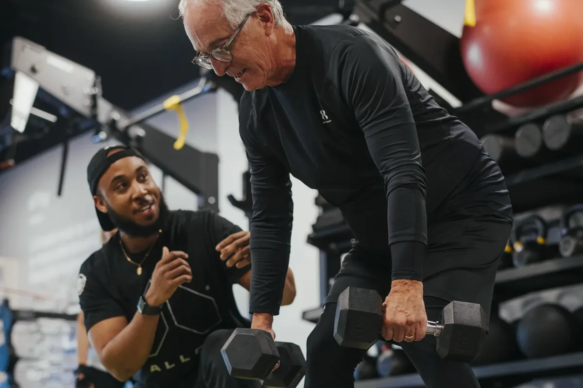A man in his sixties rows a pair of dumbbells while an Alloy coach watches his form from a crouch