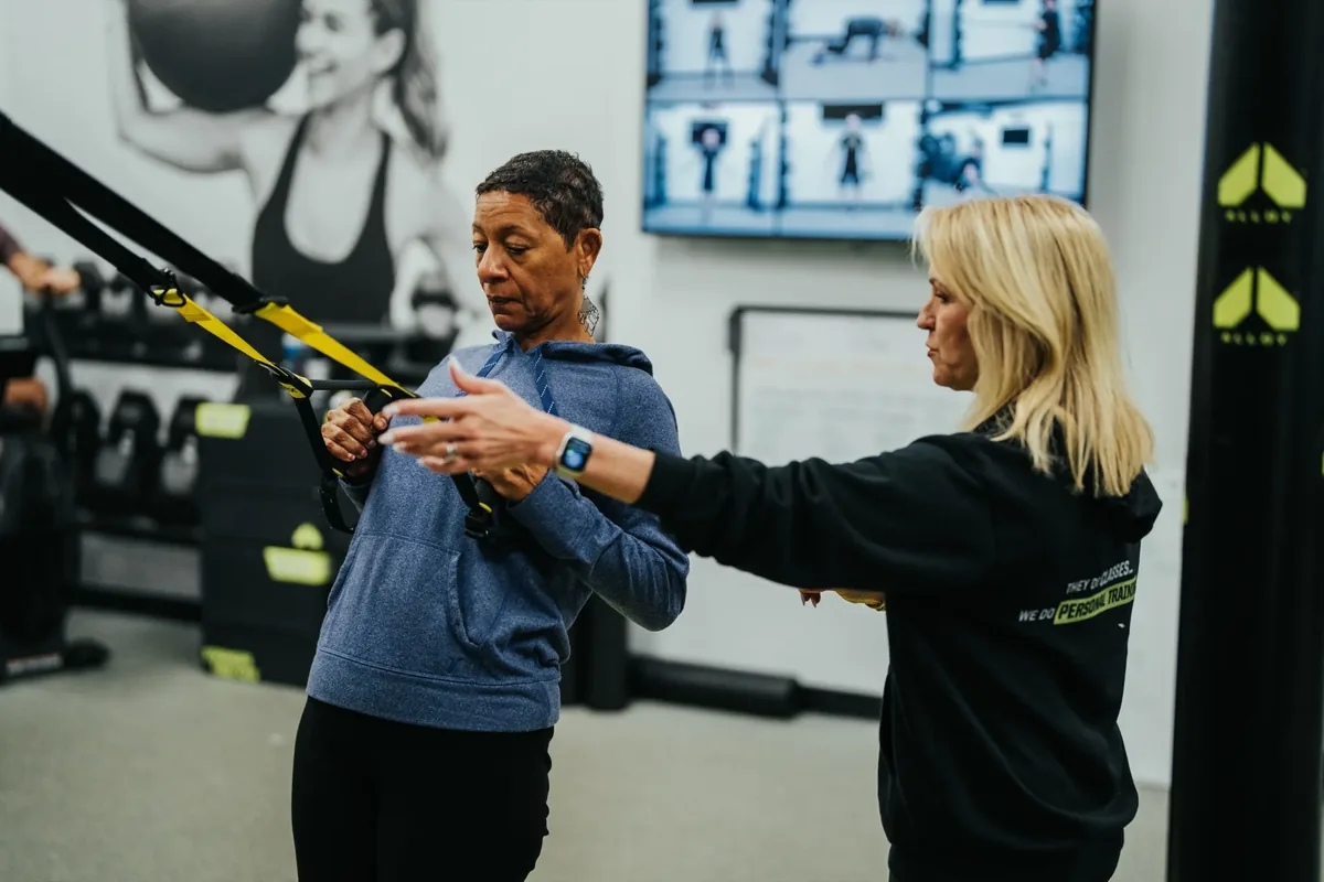 A woman in her sixties trains on suspension straps while an Alloy coach guides her arm position