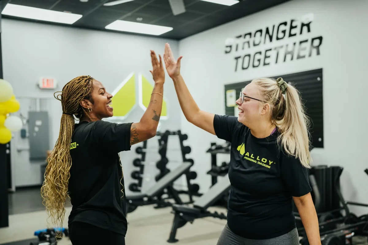 Two women in Alloy shirts share a high-five under a Stronger Together wall sign at an Alloy Personal Training studio
