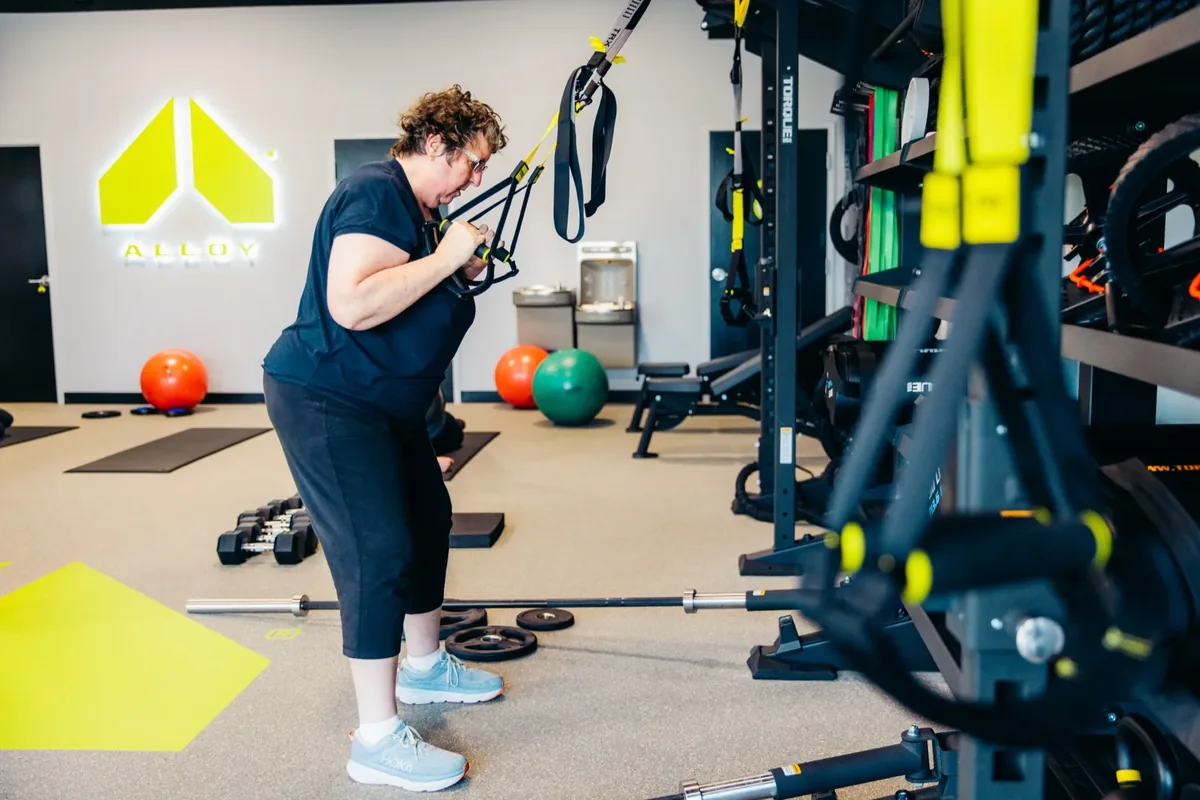 A woman trains on TRX suspension straps beside a rack on the floor of an Alloy Personal Training studio